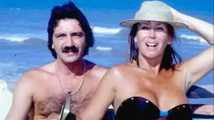 Moria Casán y su pareja posan sonrientes en traje de baño en la orilla del mar.