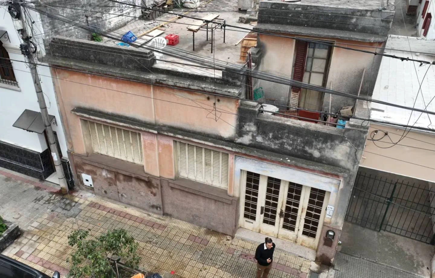 Un hombre parado en la vereda mira su celular frente a una casa antigua de persianas cerradas vista desde arriba.