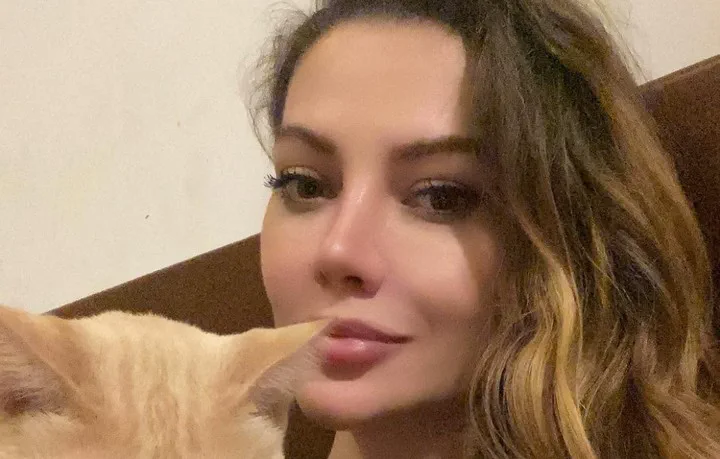 Una mujer en primer plano sonríe junto a la cabeza de un gato color claro.