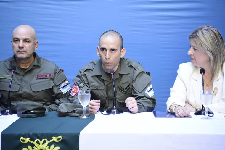 Dos hombres con uniforme militar y una mujer de saco claro están sentados frente a micrófonos.