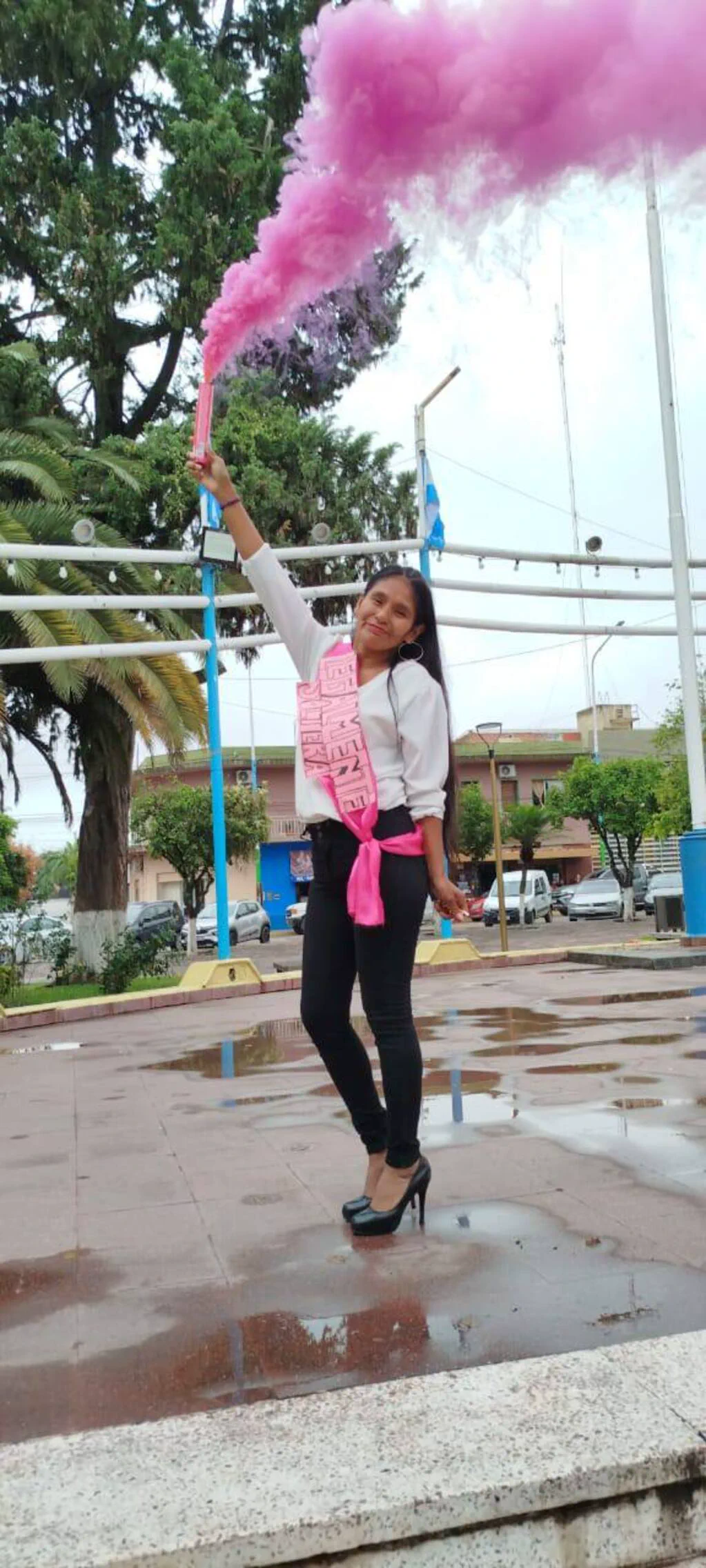 Una chica con banda rosa sostiene una bengala de humo fucsia en medio de una plaza mojada.
