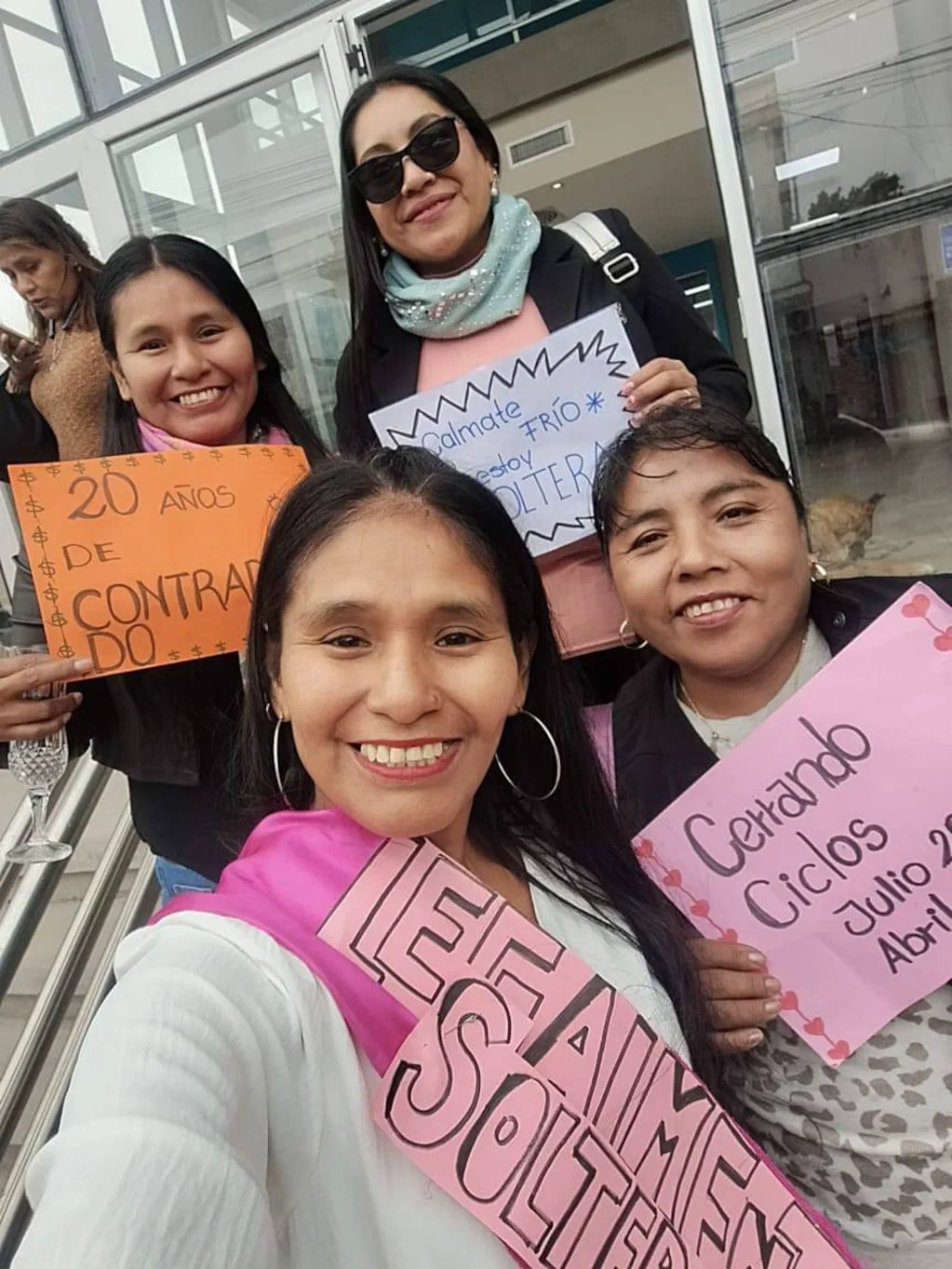 Cuatro mujeres sonríen en una escalera sosteniendo carteles manuscritos y una banda de papel.