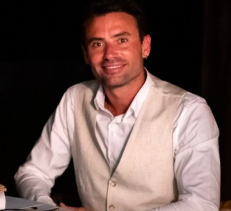 Un hombre sonriente de camisa blanca y chaleco claro esta sentado frente a un fondo oscuro.