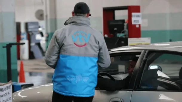 Un operario con campera de la VTV de espaldas habla con el conductor de un auto en una planta de verificación.