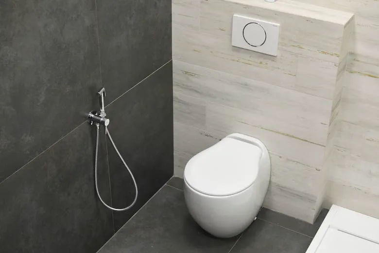 Un inodoro blanco moderno y una duchita higiénica colgada en la pared revestida con cerámicos oscuros.