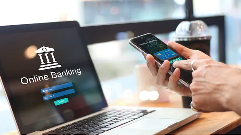 Una persona usando una aplicación bancaria en un celular frente a una notebook abierta.