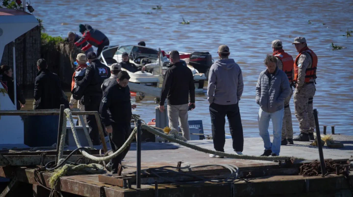 Varios hombres y efectivos de seguridad están parados sobre un muelle metálico frente a embarcaciones en el agua.