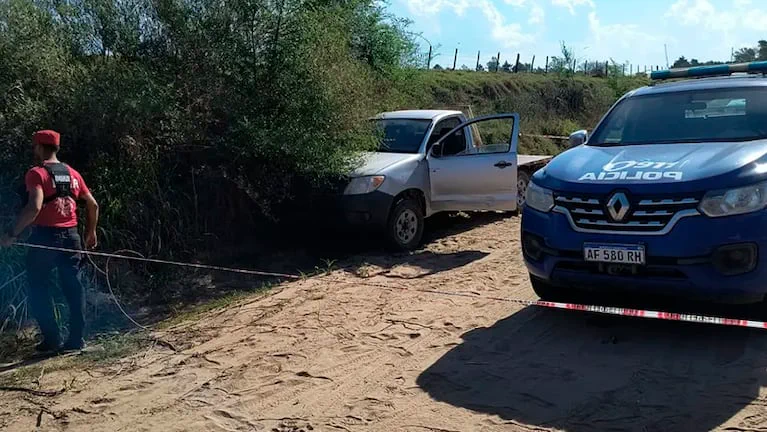 Un patrullero azul y una camioneta gris permanecen detenidos en un camino de tierra delimitado con cinta.