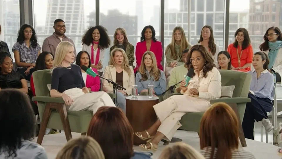 Oprah Winfrey y otra mujer conversan sentadas en un panel frente a un público en una sala luminosa.