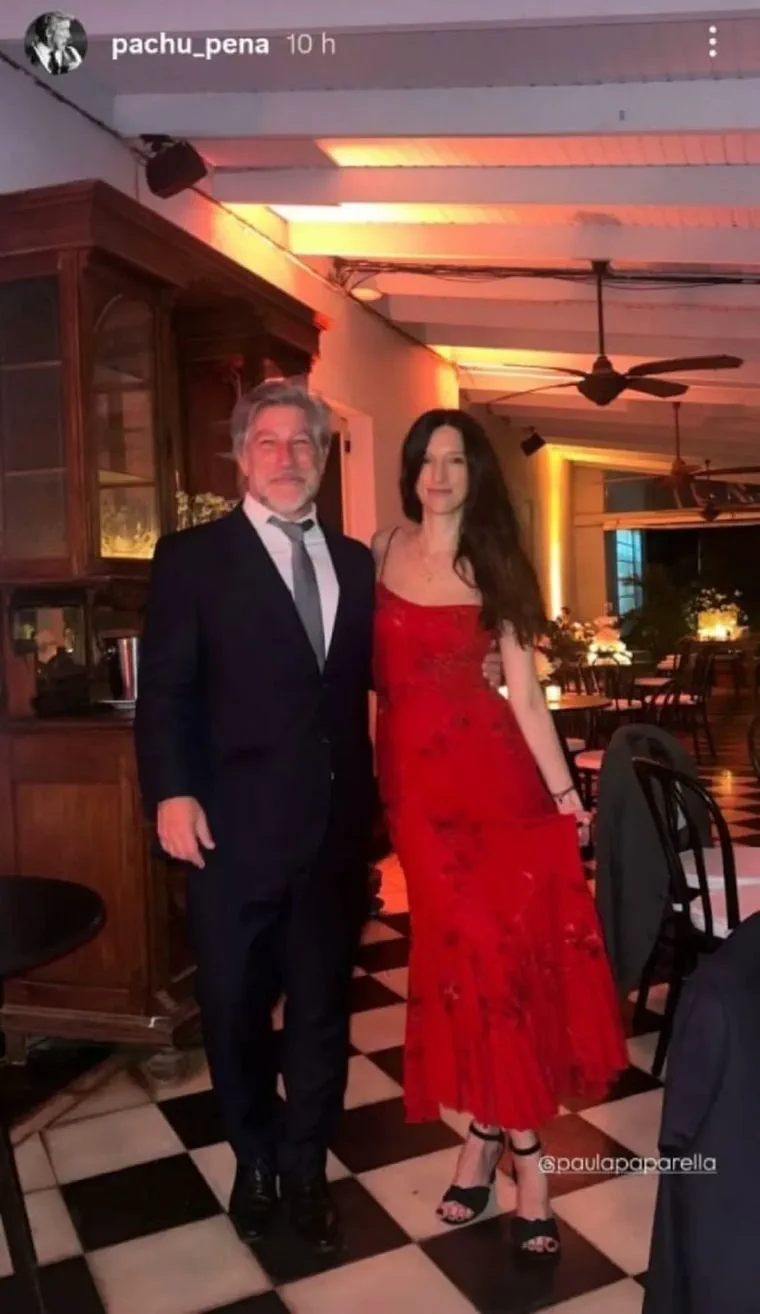 Pachu Peña de traje oscuro junto a una mujer con vestido largo rojo en un salón.
