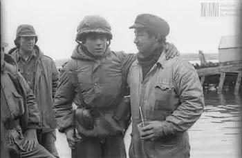 Un soldado y un hombre con gorra y campera conversan al aire libre cerca del agua.