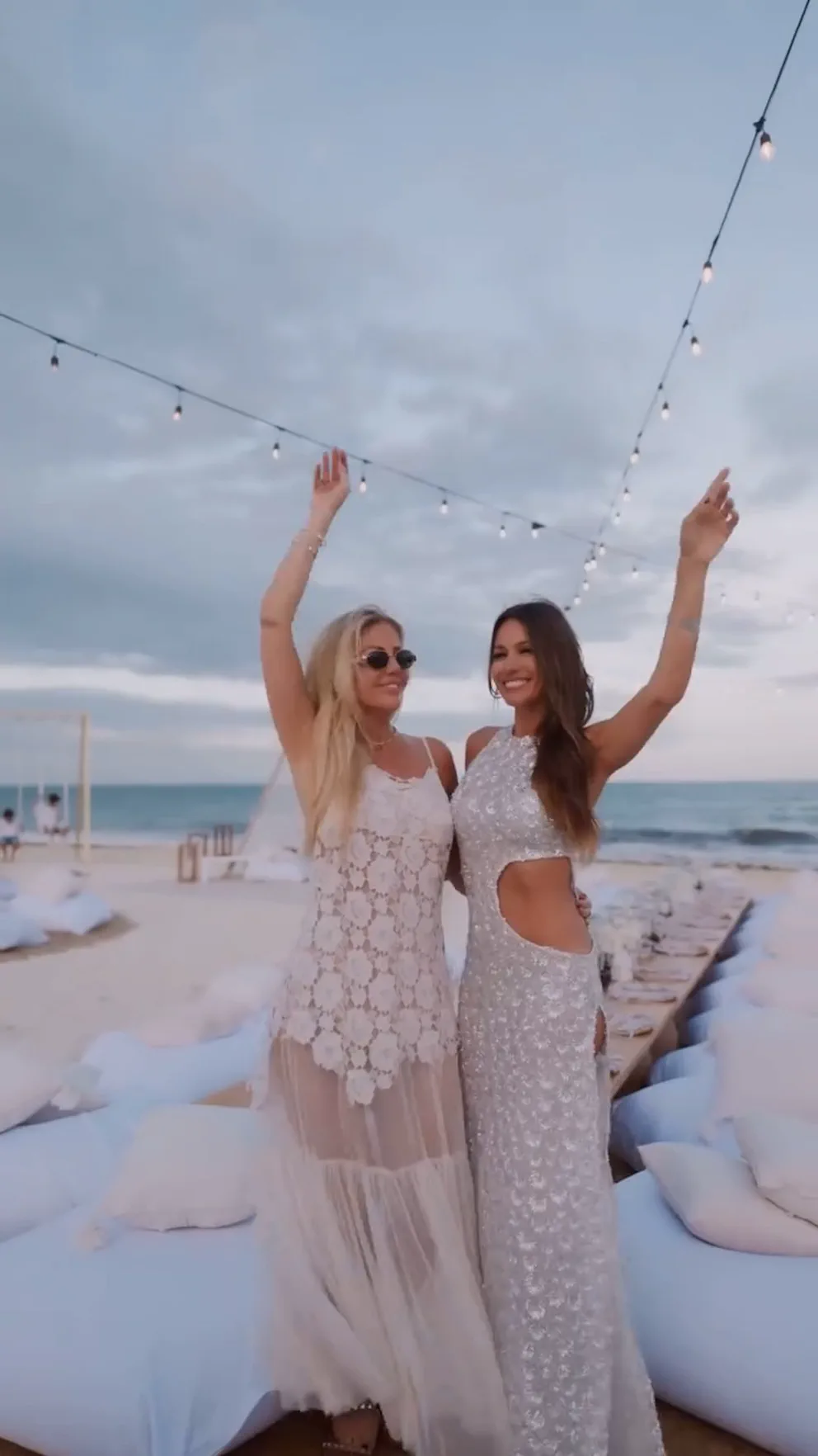 Pampita y otra mujer con vestidos blancos sonríen con los brazos en alto en una playa.