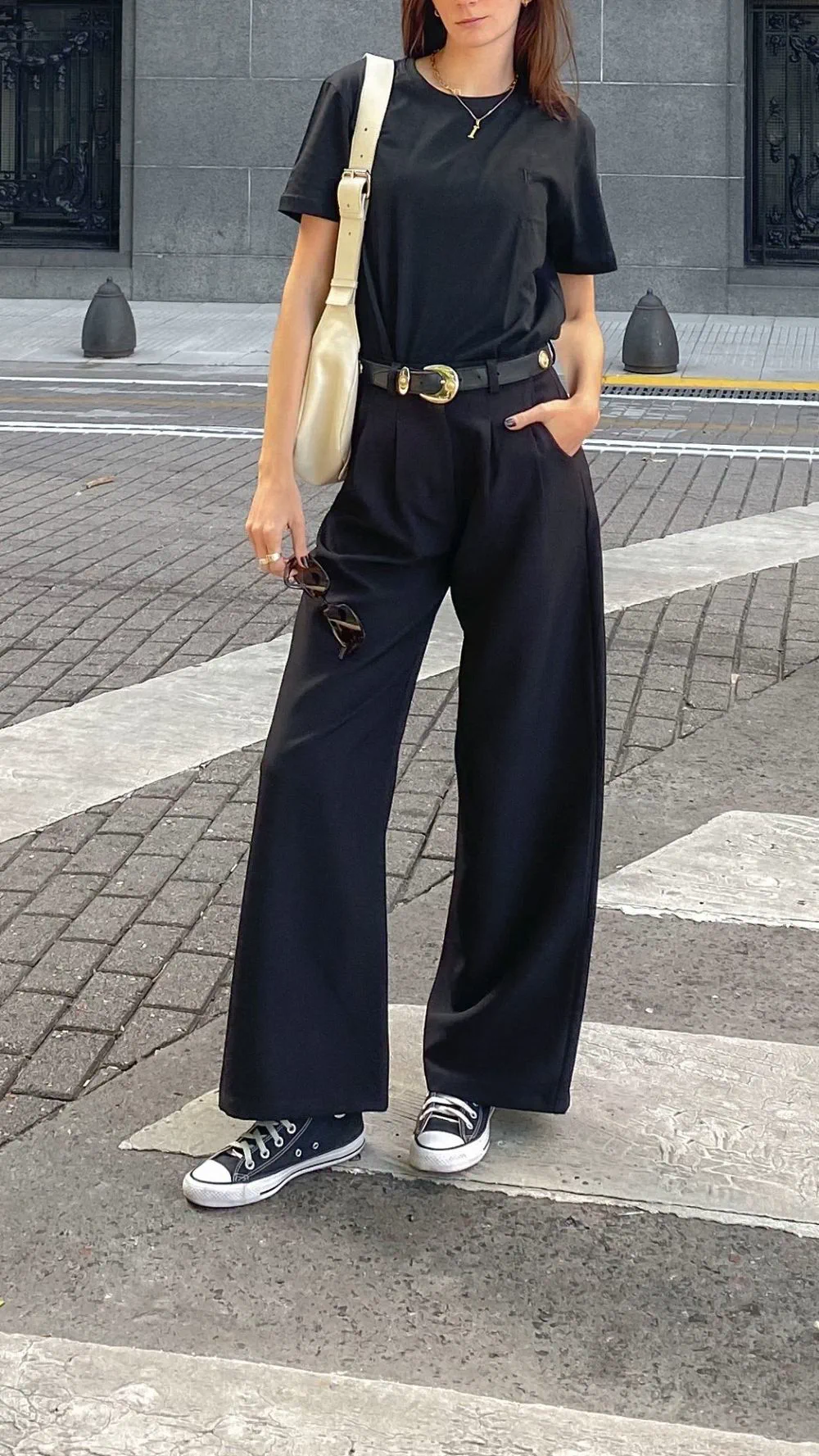 Una mujer parada sobre una senda peatonal viste remera y pantalón ancho negros, zapatillas y cartera clara.