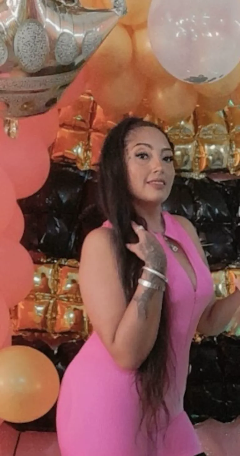 Una mujer con vestido rosa y tatuajes en el brazo posa delante de una pared decorada con globos.
