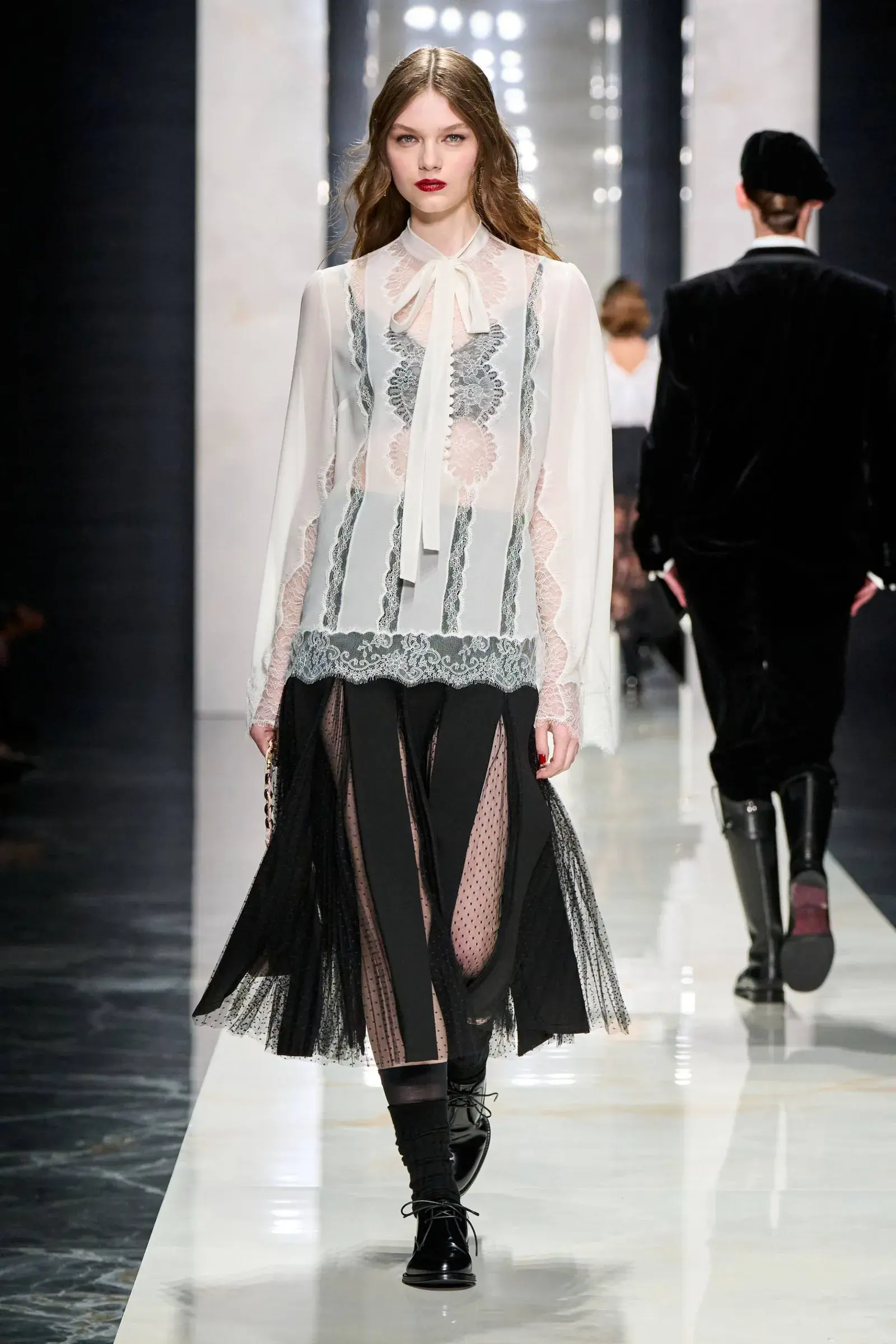 Modelo desfilando en una pasarela con blusa blanca de encaje y pollera negra con transparencias.