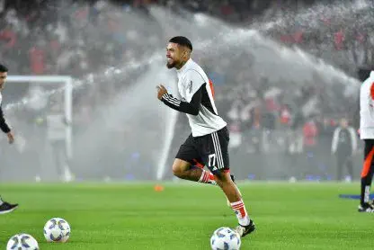 El futbolista Paulo Diaz corre y sonrie sobre el cesped de una cancha de futbol con aspersores encendidos de fondo.
