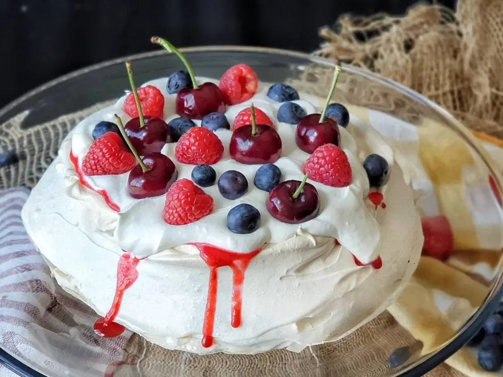 Una torta pavlova cubierta de crema batida, salsa roja, frambuesas, cerezas y arandanos sobre una fuente de vidrio.