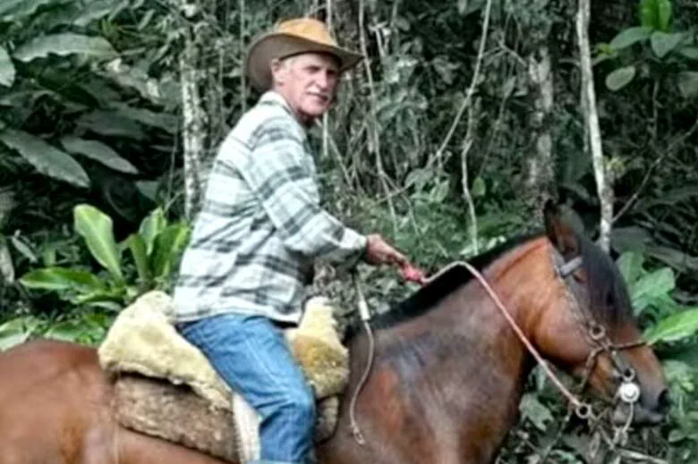 Un hombre con sombrero de campo y camisa a cuadros monta un caballo castaño en medio del monte.