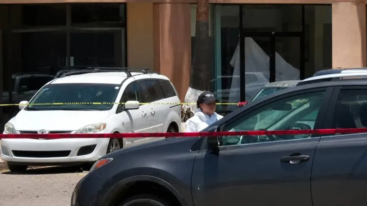 Una persona con gorra y campera blanca parada detrás de una cinta de peligro entre dos autos estacionados.