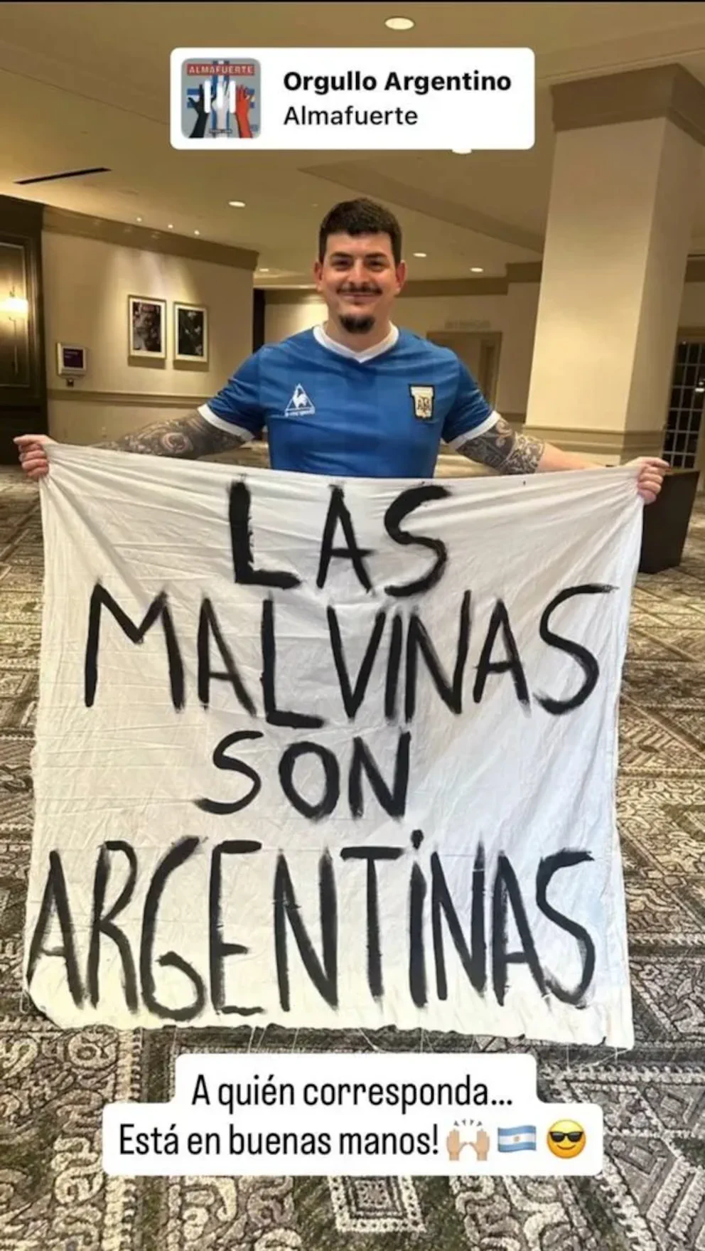 Hombre con camiseta de la selección argentina posa en un salón sosteniendo una tela con consigna sobre Malvinas.