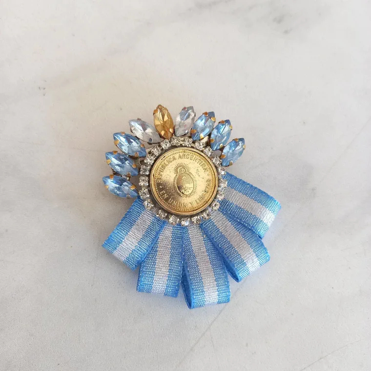 Un prendedor patrio con una moneda argentina central rodeada de piedras facetadas y una cinta celeste y blanca plegada.