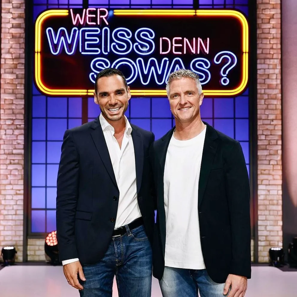 Dos hombres sonrientes parados de pie posan adelante de un letrero luminoso de televisión.