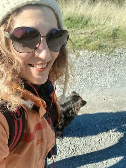 Una mujer con anteojos de sol y gorro de lana sonríe en un camino de ripio junto a un perro negro.