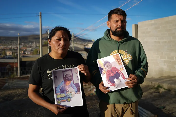 Una mujer y un hombre sostienen fotos impresas de un nene al aire libre en un terreno descampado.