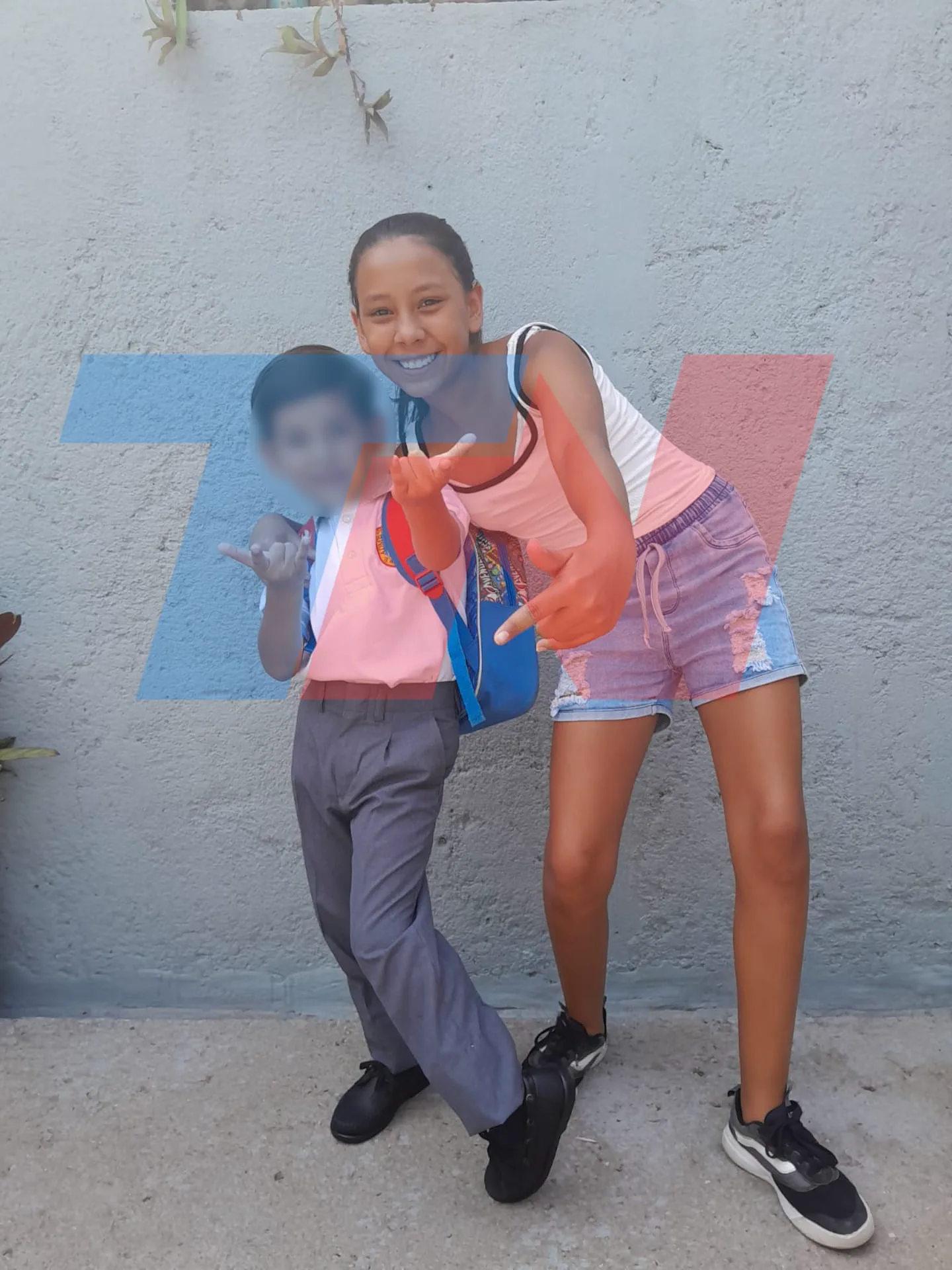 Una chica con remera rosa y short de jean posa junto a un nene de uniforme con la cara borrosa frente a una pared.