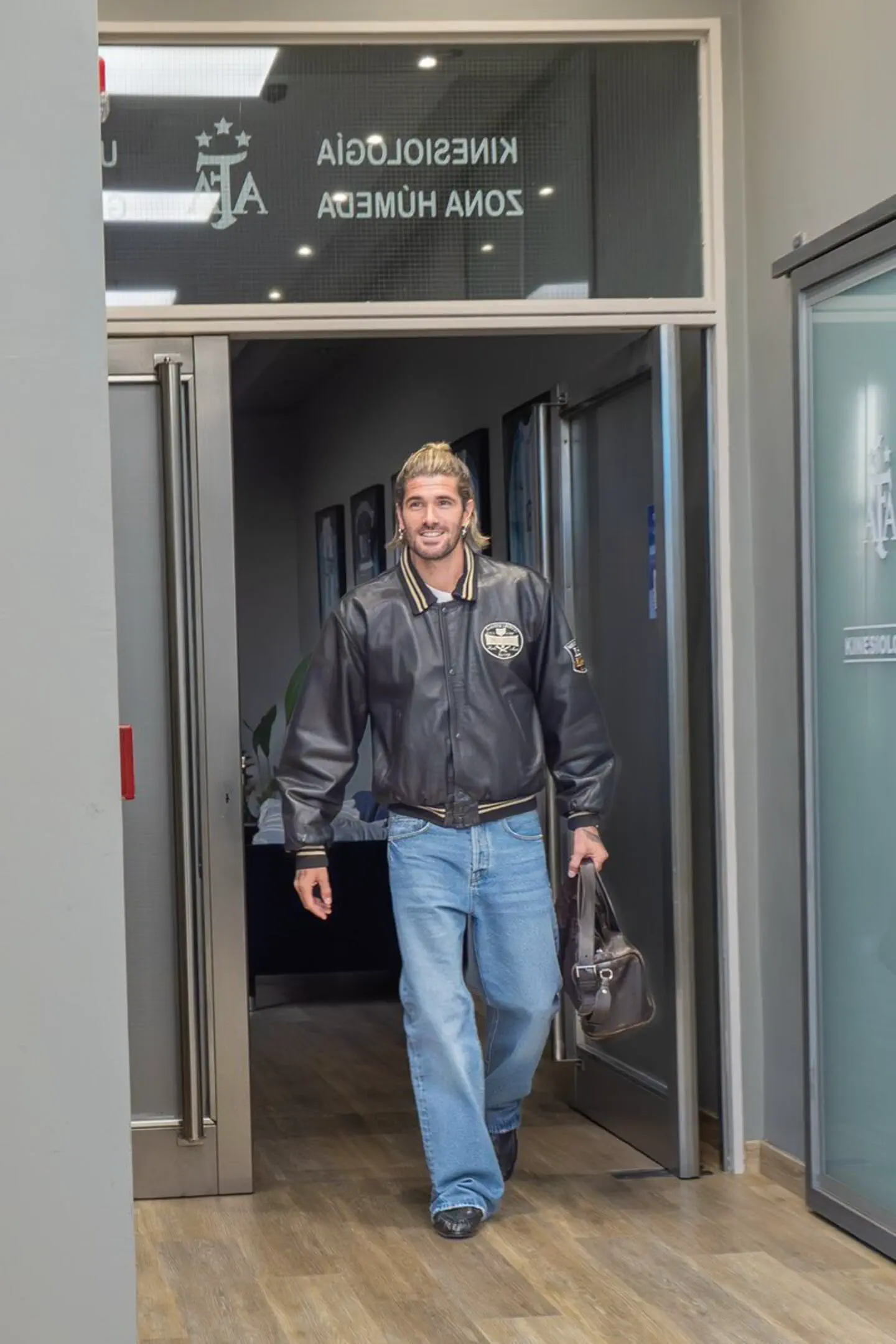 Rodrigo De Paul entrando por una puerta con campera de cuero, jean ancho y bolso de mano.