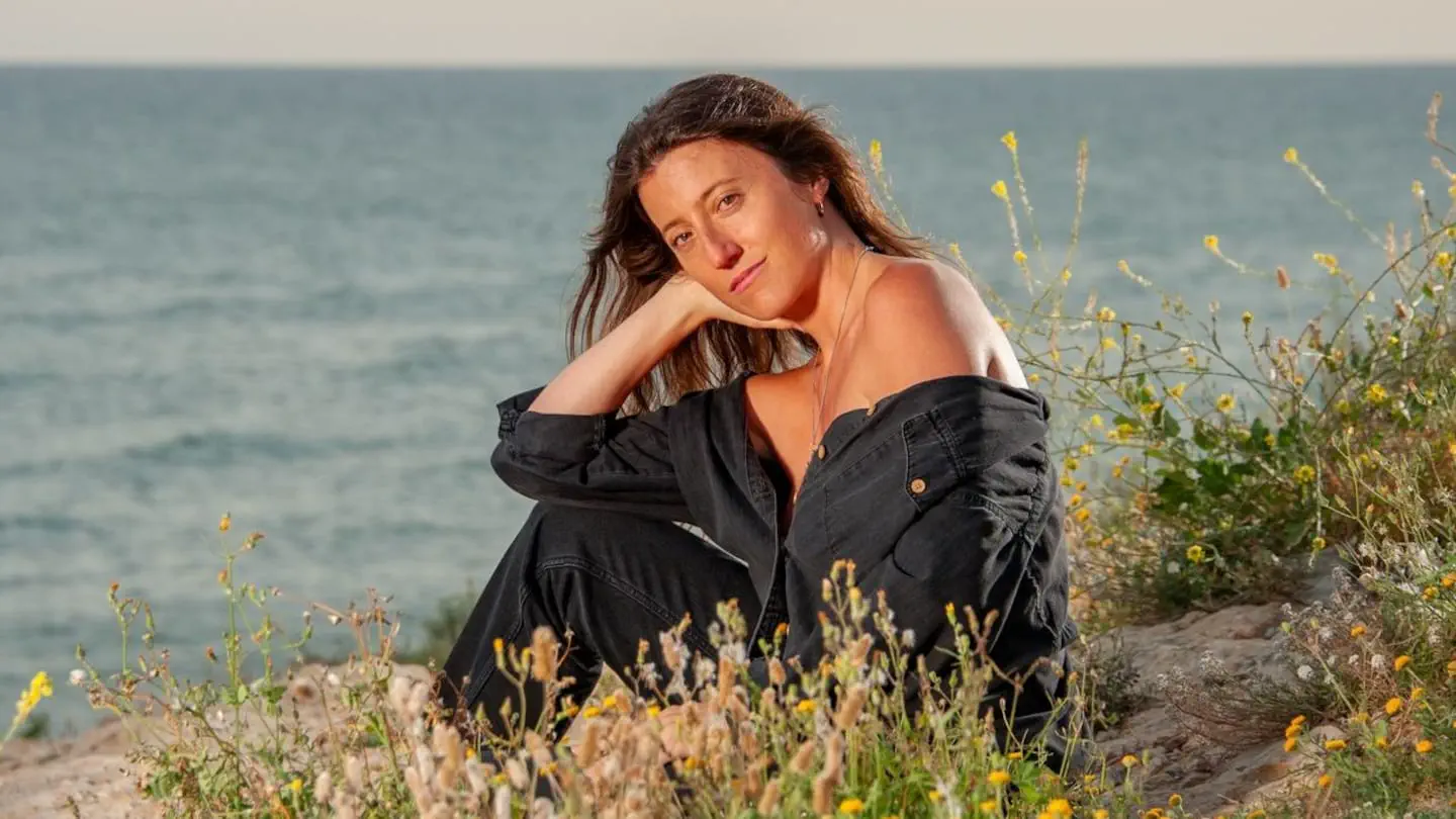 Una mujer con campera negra descasada de hombros posa sentada entre plantas silvestres frente al mar.