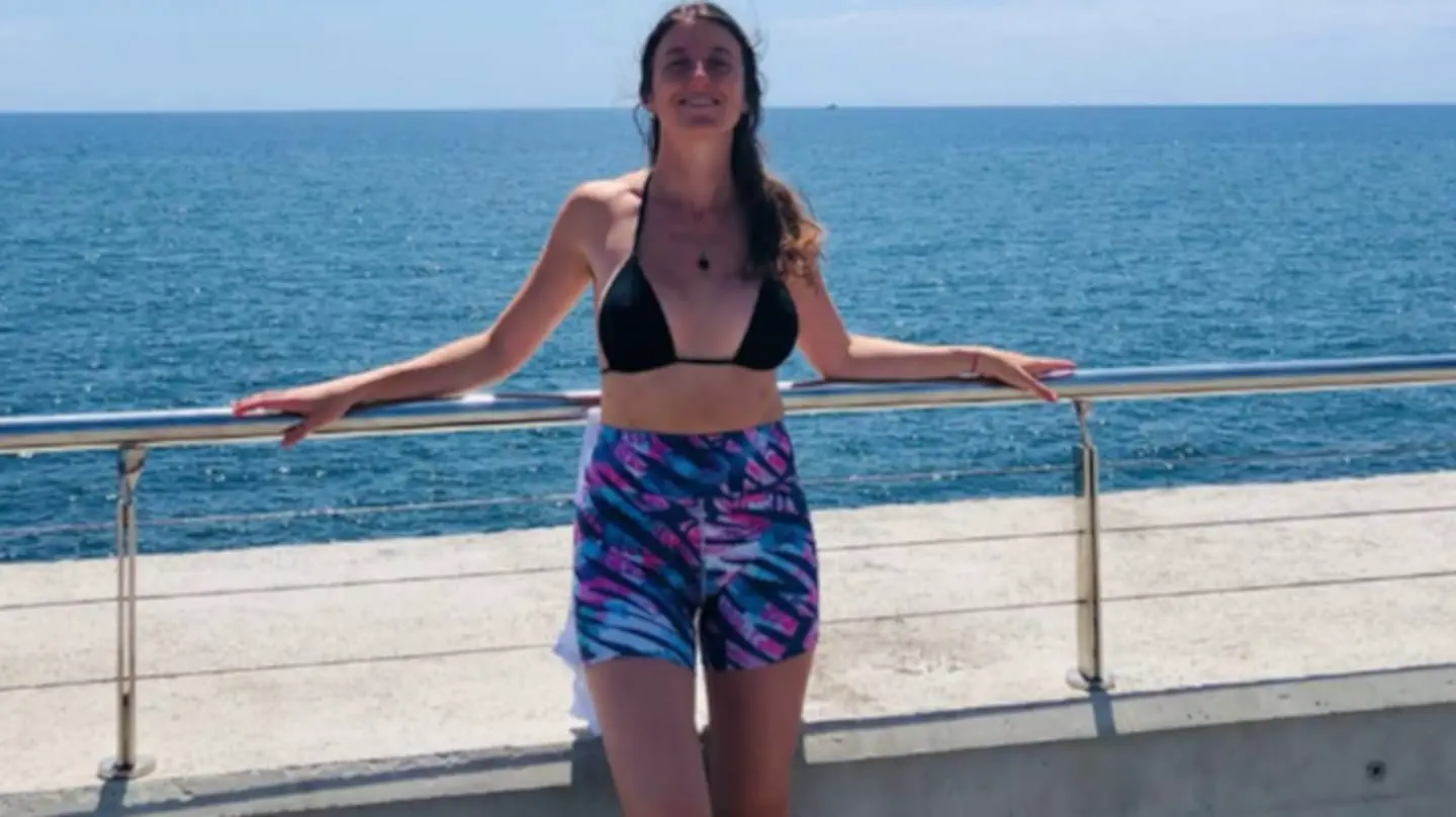 Una mujer en bikini y calzas cortas sonrie apoyada en una baranda frente al mar.