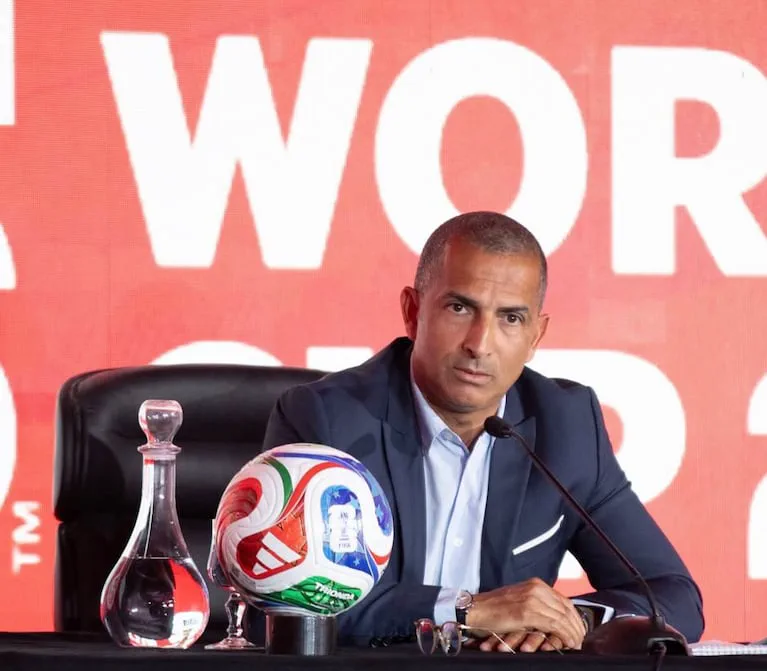 Sabri Lamouchi sentado en una mesa de conferencia con un microfono, una pelota y una botella de vidrio al frente.