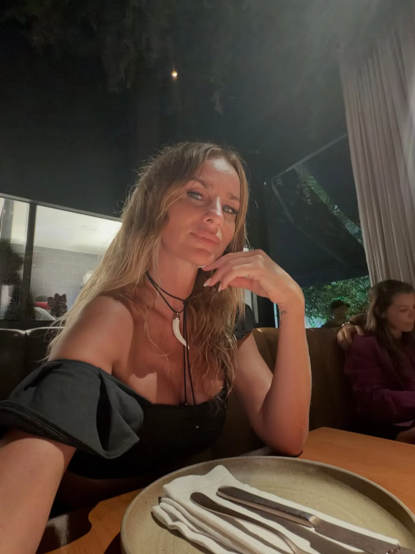 Una mujer rubia con vestido negro escotado y dije blanco al cuello posa sentada a la mesa de un restaurante.