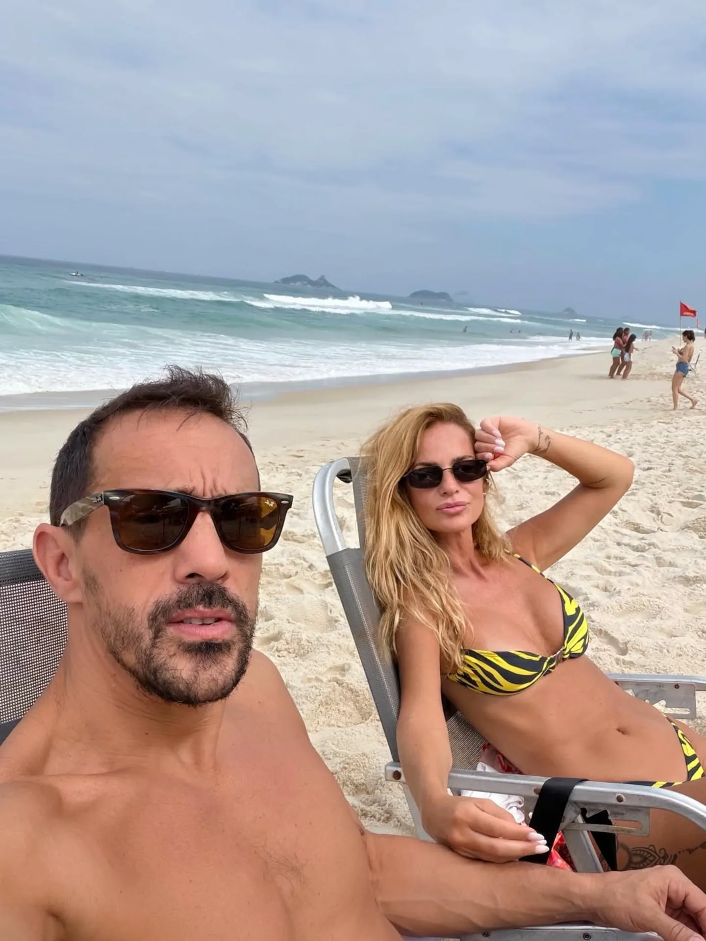Un hombre y una mujer en reposera toman sol con anteojos oscuros en la orilla de una playa concurrida.