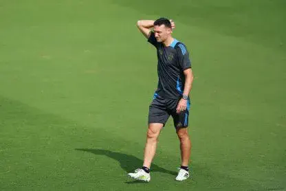 Lionel Scaloni camina sobre el pasto de una cancha con una mano en la cabeza, vestido con indumentaria deportiva.
