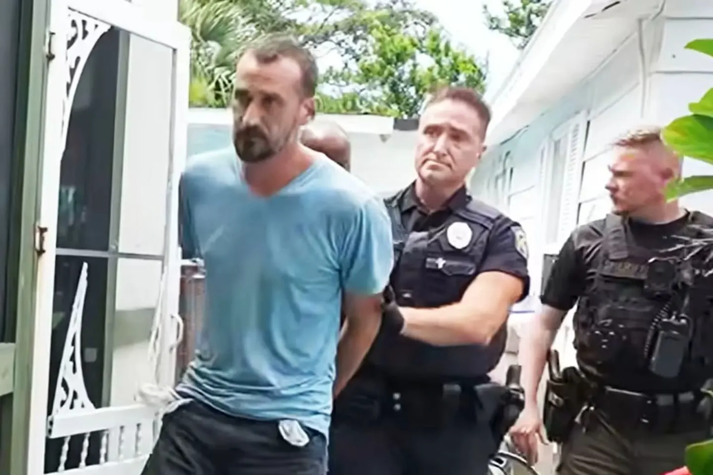 Un hombre esposado con remera celeste camina escoltado por dos policías al lado de una vivienda.