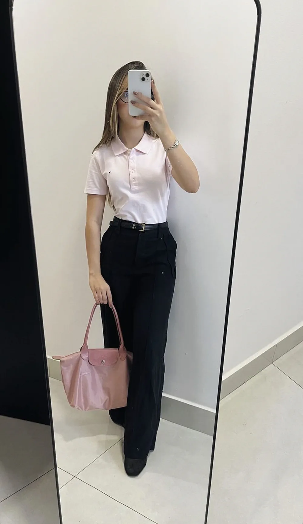 Una joven se saca una selfie frente a un espejo llevando una chomba rosa claro, pantalón negro y bolso rosa.