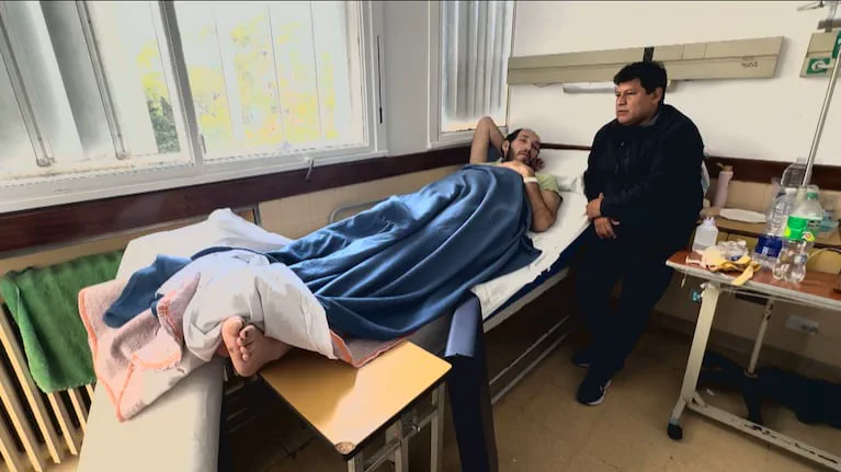Un hombre acostado en una cama de hospital tapado con una manta azul mientras otro hombre está sentado a su lado.