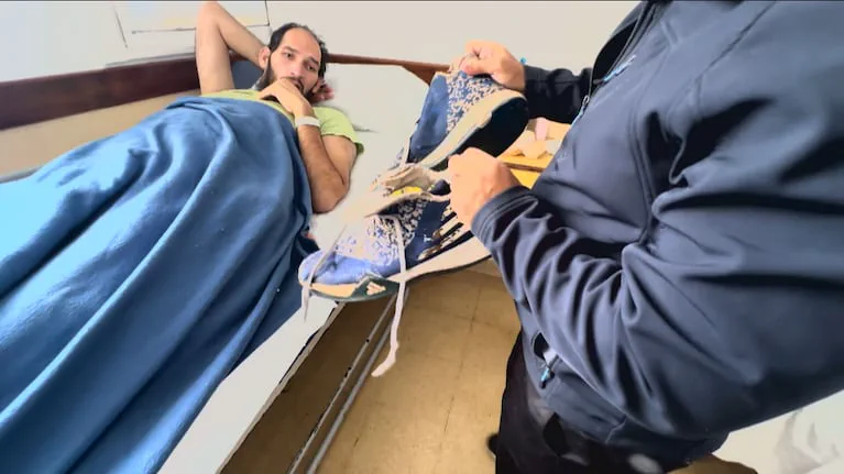 Un hombre parado sosteniendo un par de zapatillas azules frente a otro que está recostado en una cama de hospital.
