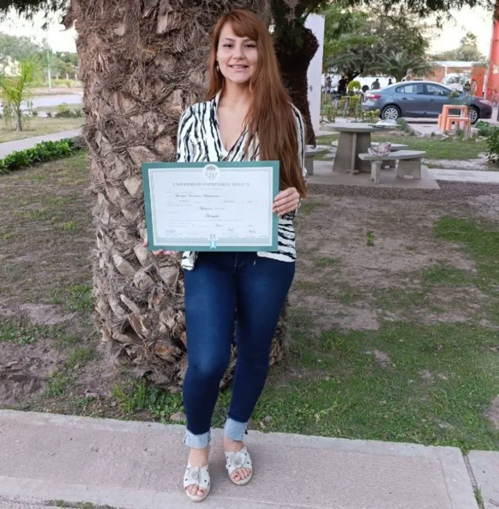 Shadya Altamirano sonríe sosteniendo su diploma universitario de abogada junto a una palmera.