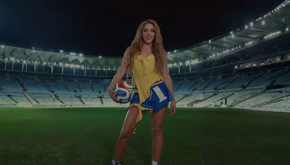 Shakira posando de pie con una pelota de fútbol bajo el brazo en el césped de un estadio iluminado.