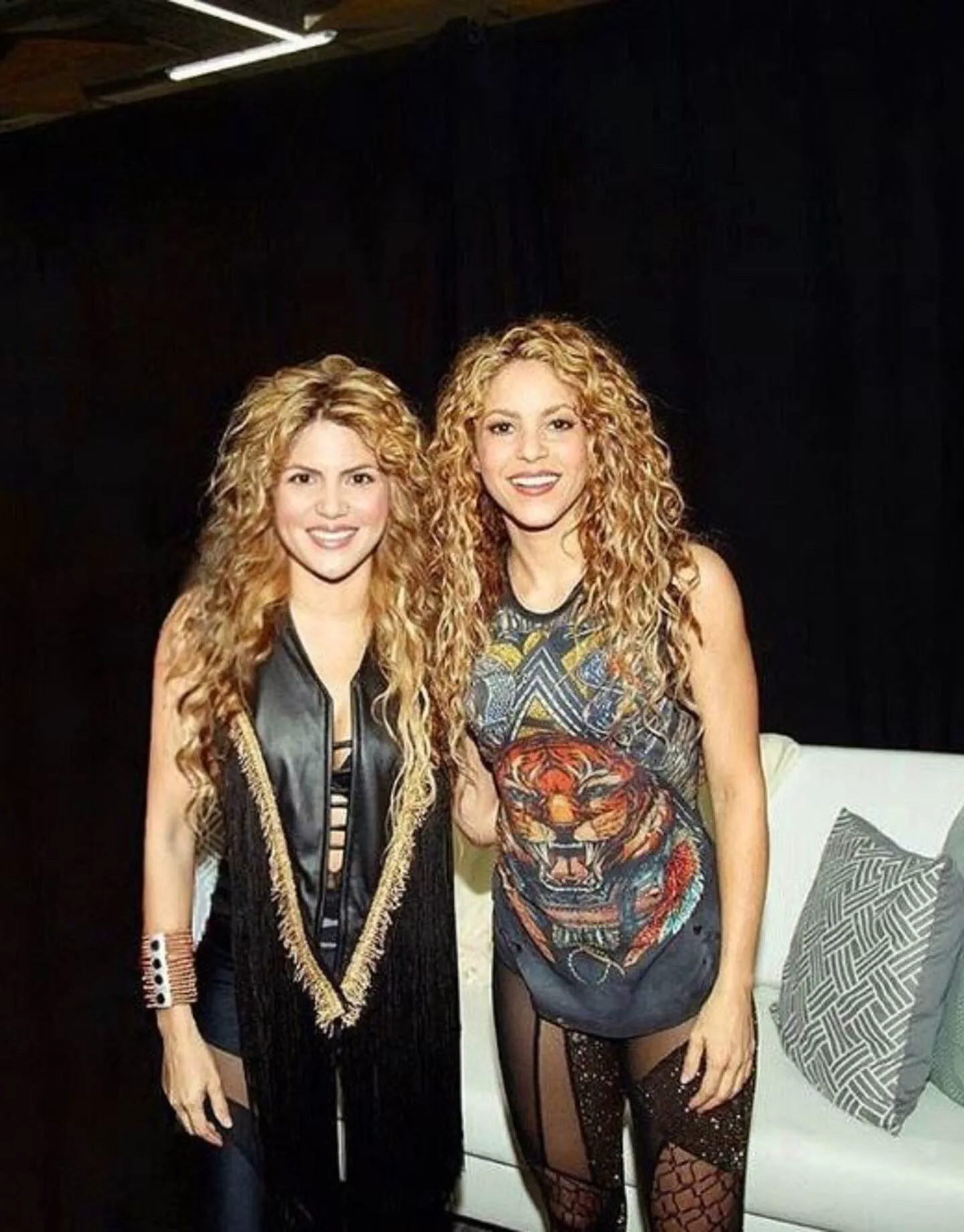 La cantante Shakira posa sonriendo parada junto a su doble o imitadora en un espacio interior.