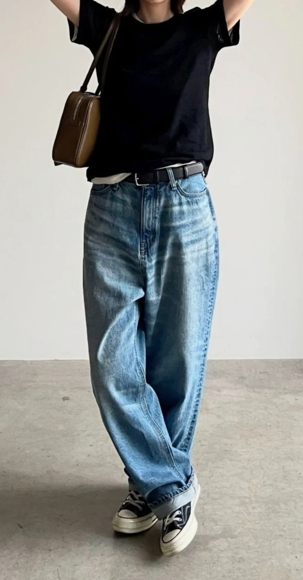 Una persona con remera negra, jean holgado y zapatillas de lona posa con los brazos en alto llevando un bolso.
