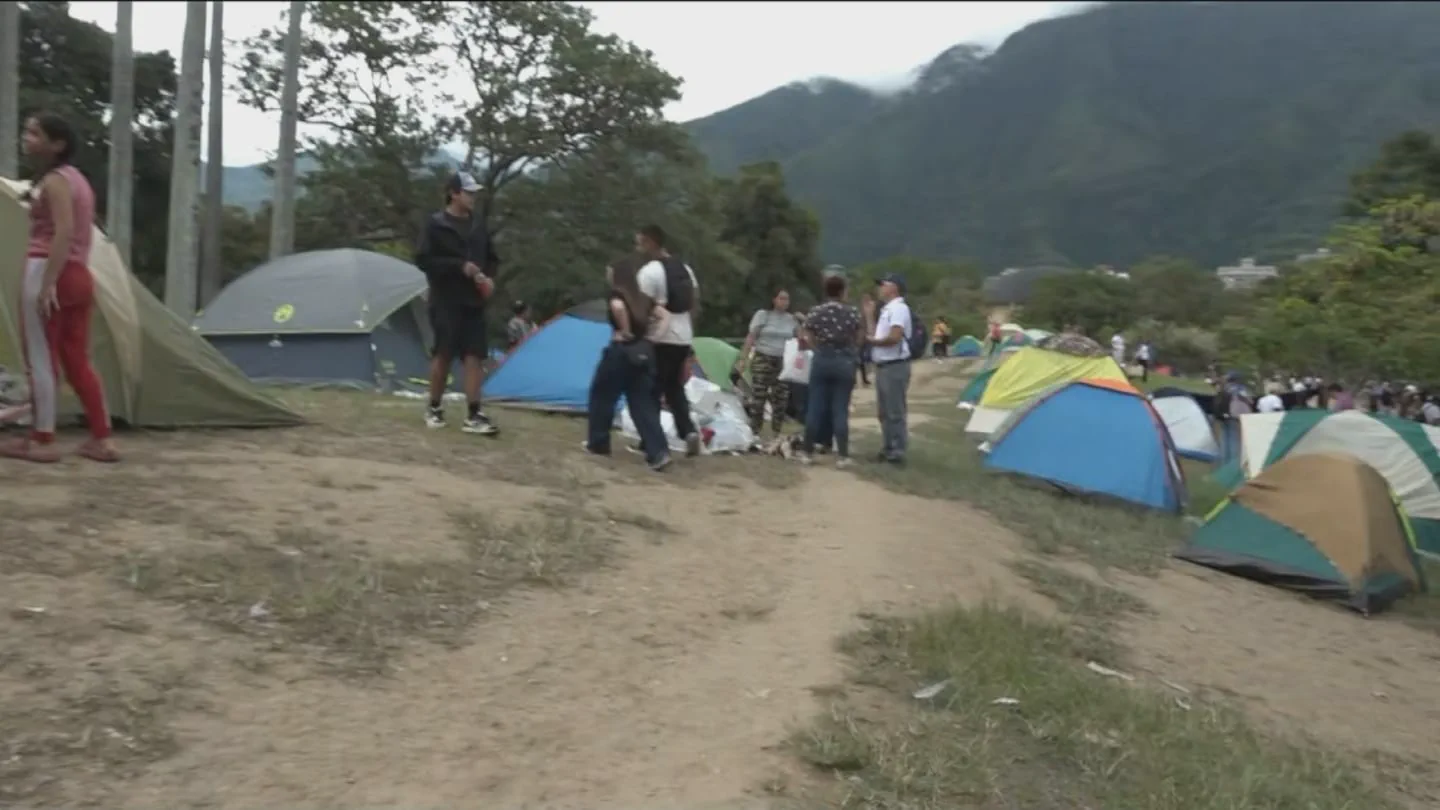 Varias personas caminan y conversan en un campamento al aire libre rodeado de carpas y montañas.
