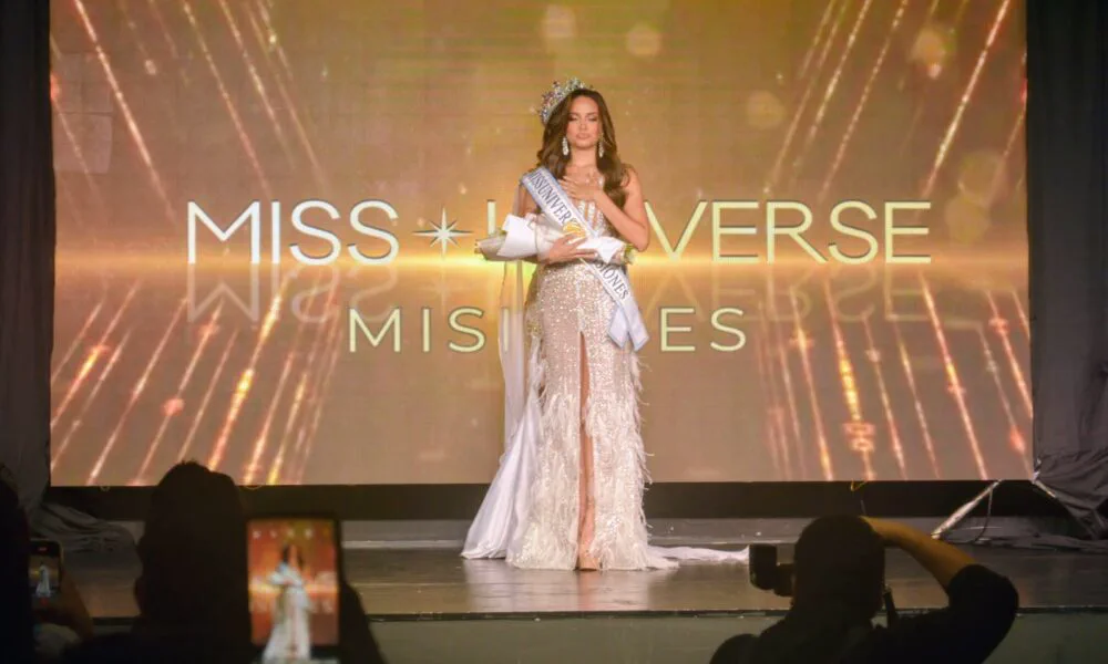 Una mujer con corona, vestido largo brillante y banda de concurso posa sobre un escenario.