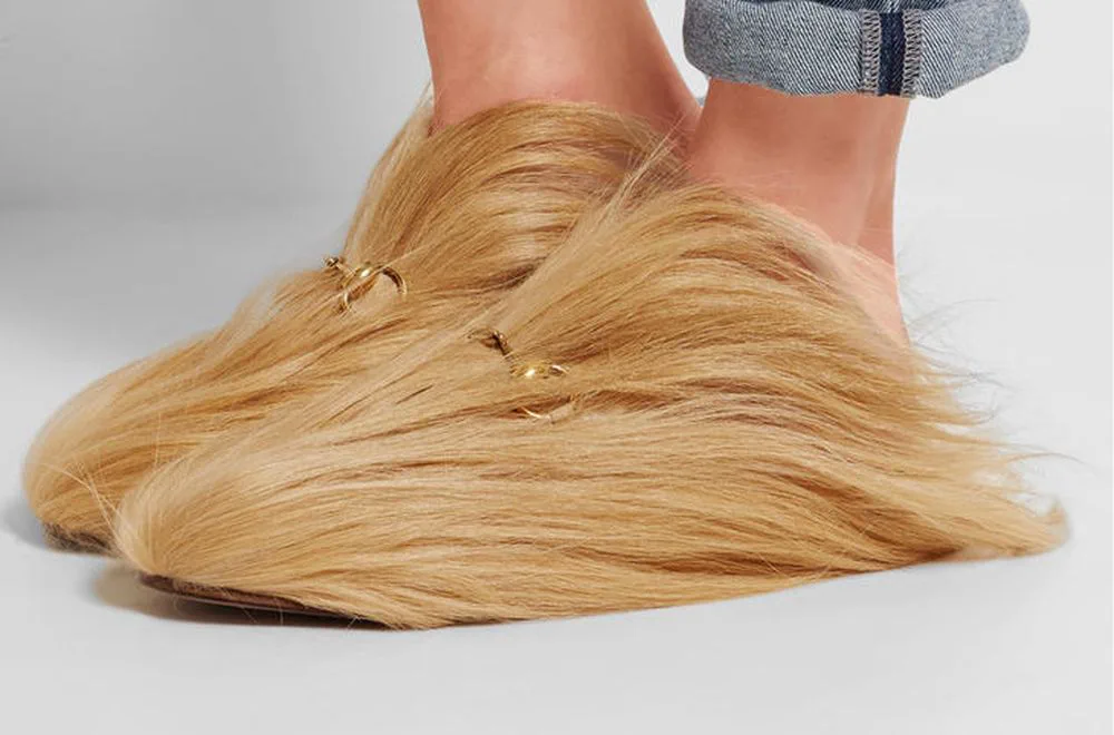Pies de una persona calzando pantuflas o zapatos cubiertos por completo de pelo rubio con hebillas doradas.