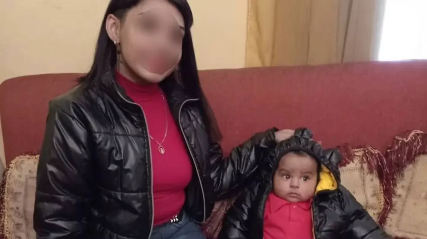 Una mujer con el rostro difuminado y un bebé con capucha negra están sentados en un sillón rojo.