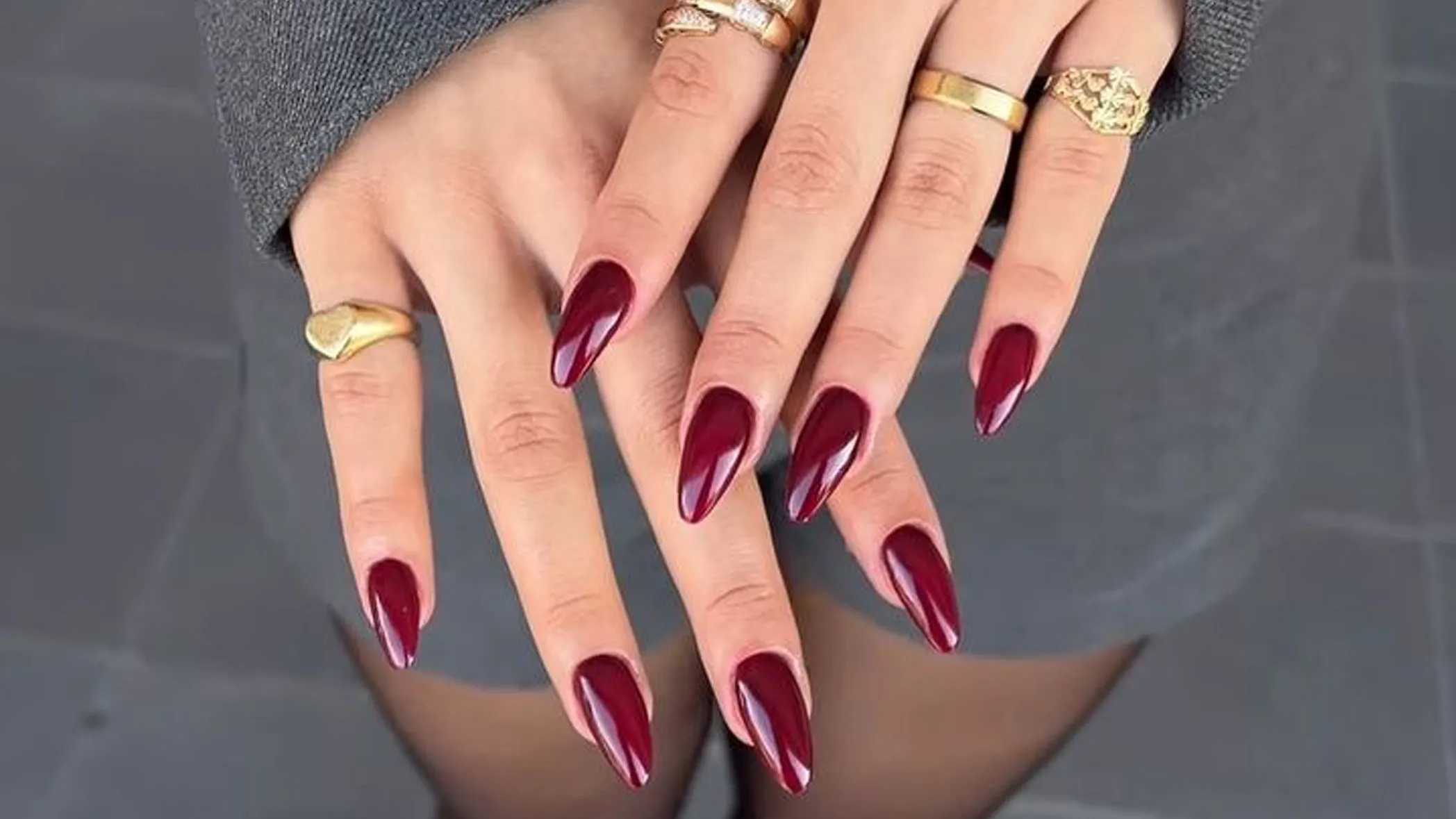 Un par de manos lucen uñas postizas largas y afiladas pintadas de bordó brillante junto a varios anillos dorados.