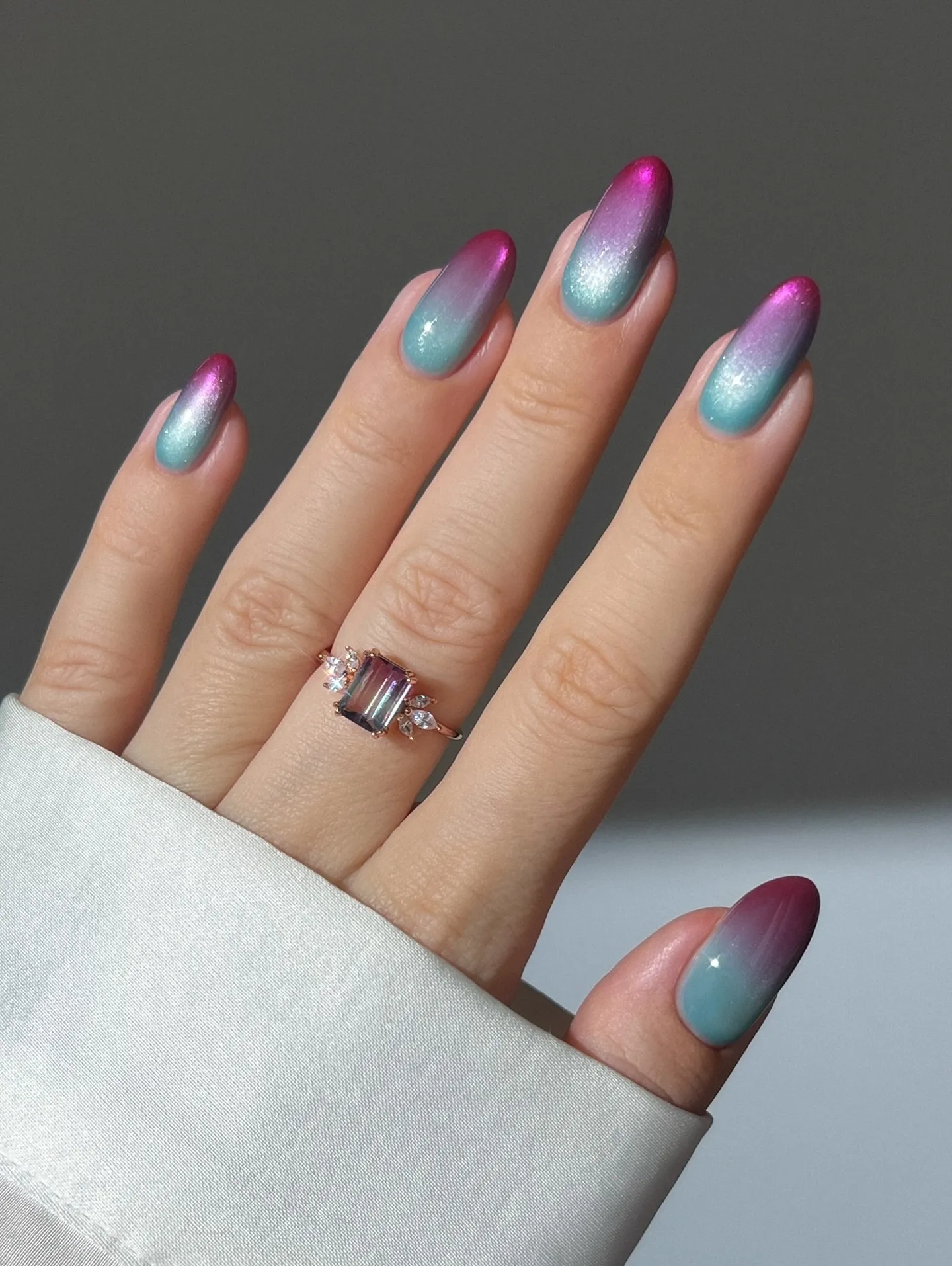 Una mano luciendo uñas pintadas en degradé brillante con tonos celestes y fucsias, y un anillo.